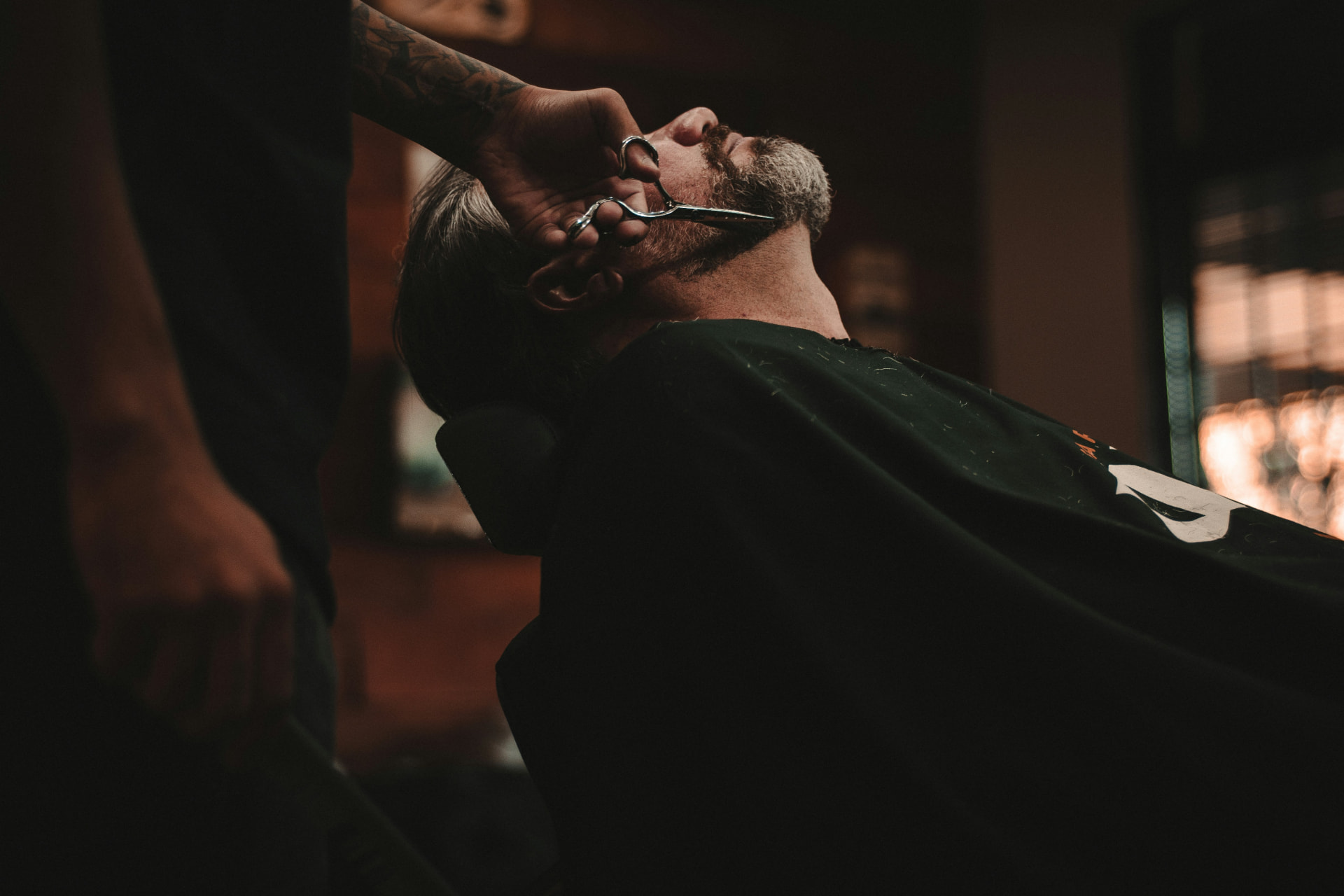 A barber trimming a client's beard with shears in a dimly lit shop